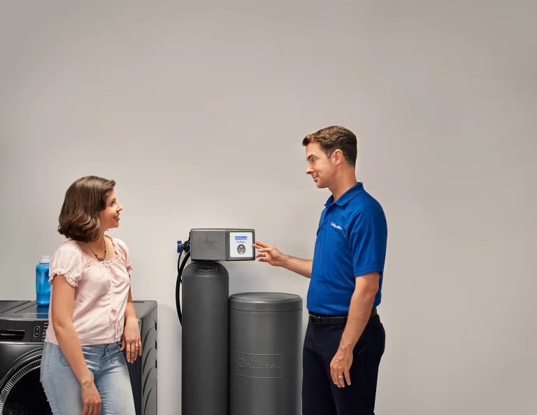 Une femme se tient à côté d'une machine à laver et d'un adoucisseur d'eau Culligan tandis qu'un technicien en polo bleu lui explique le système de filtration de l'eau en montrant son panneau de commande. L’arrière-plan est un mur uni de couleur claire.