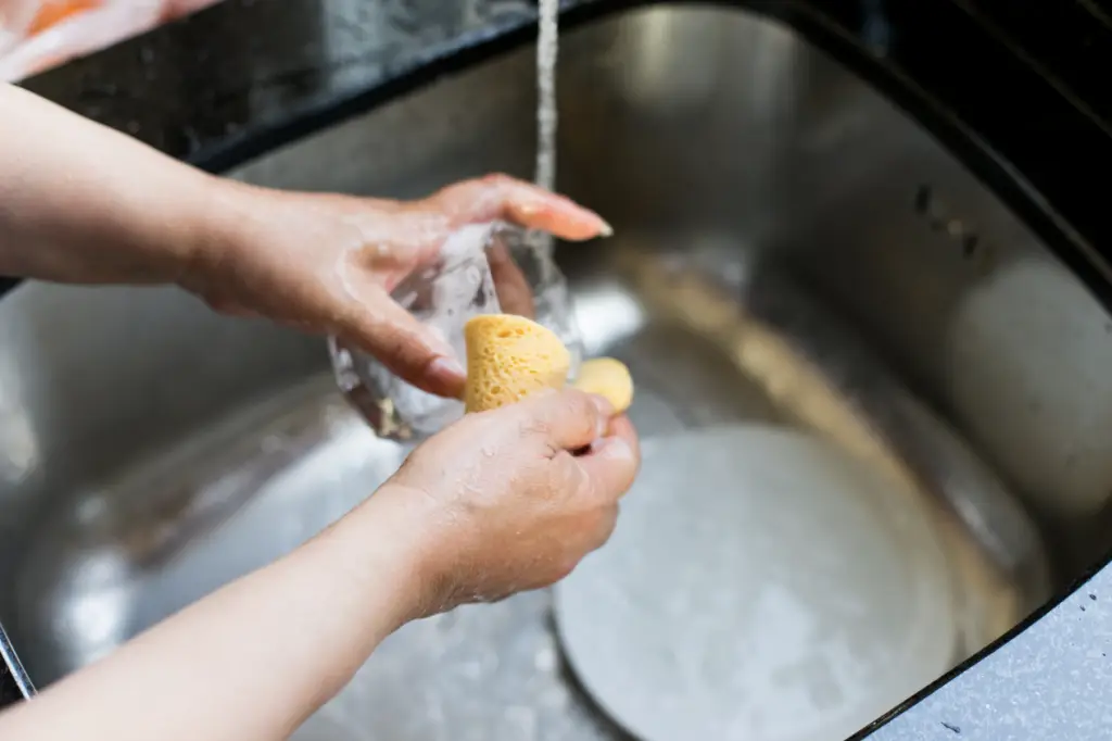 Femme qui la un verre sous l'eau d'un évier de cuisine avec une éponge jaune