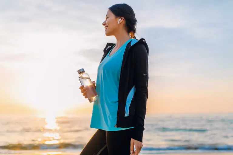 Une femme qui sourie devant l'océan avec une bouteille d'eau à la main vêtu de vêtement d'exercise